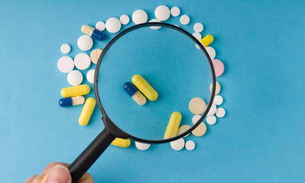 A hand holding a magnifying glass. The magnifying glass amplifies an assortment of colorful pills against a blue background.