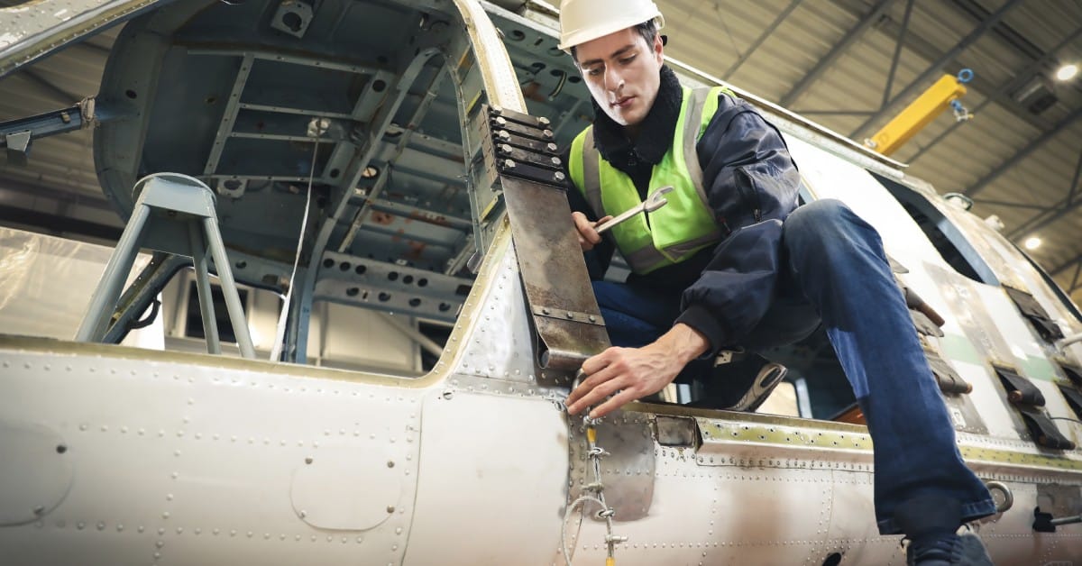 A lower view of a male factory worker in a hard hat and high-visibility vest connecting metal cables to a helicopter frame.