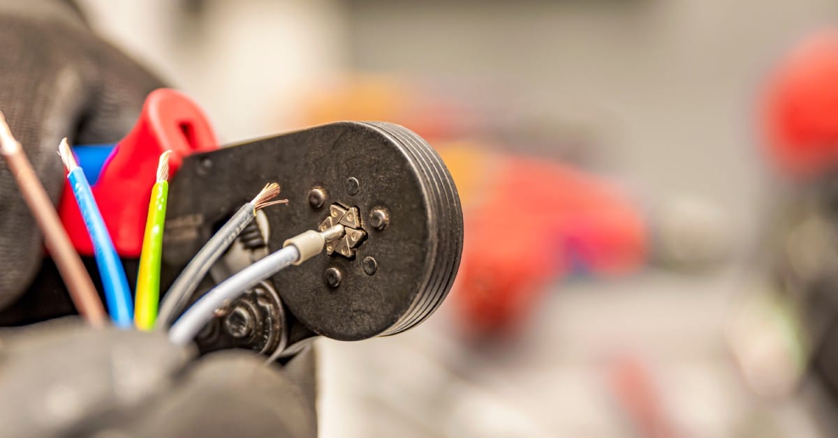 A close-up view of a handheld wire crimping tool as a hand closes the crimper on a silver wire cable.