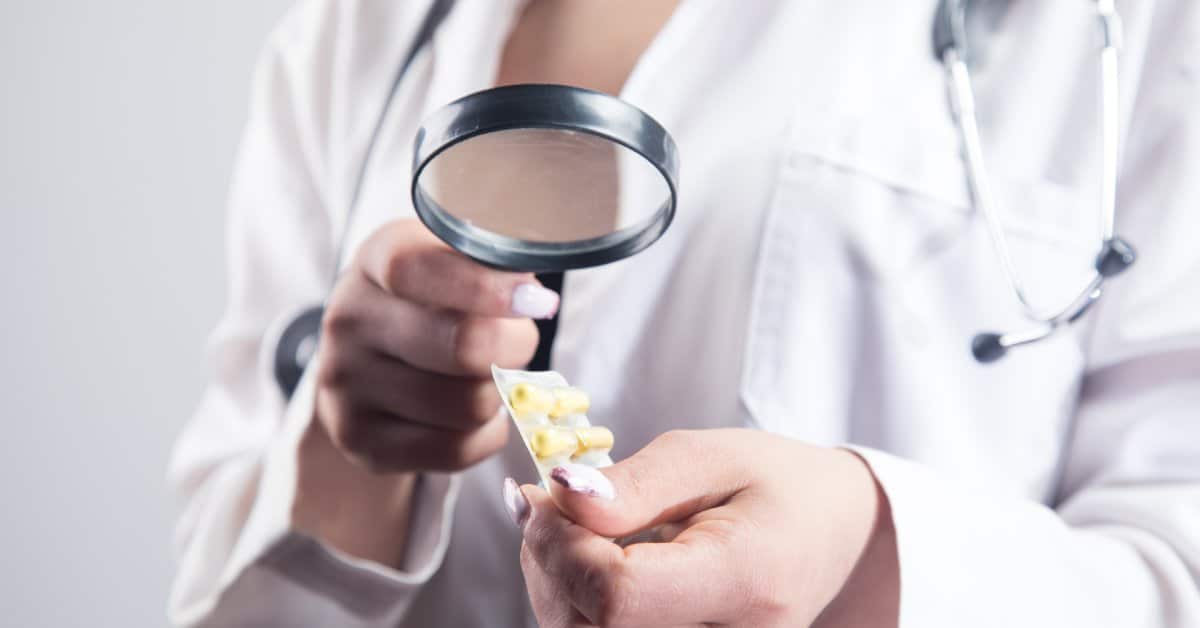 A female doctor in a white lab coat holds a package of yellow pharmaceutical pills under a magnifying glass.