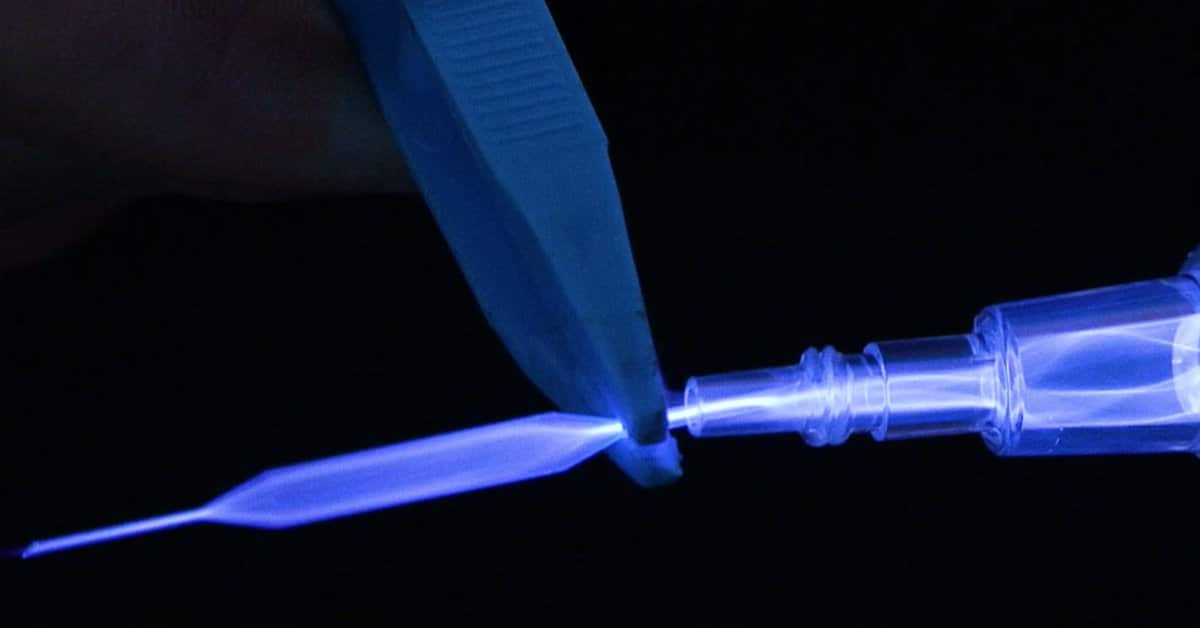 A glowing blue atmospheric plasma jet being generated from a transparent nozzle. A pair of tweezers is positioned near the plasma stream against a dark background.