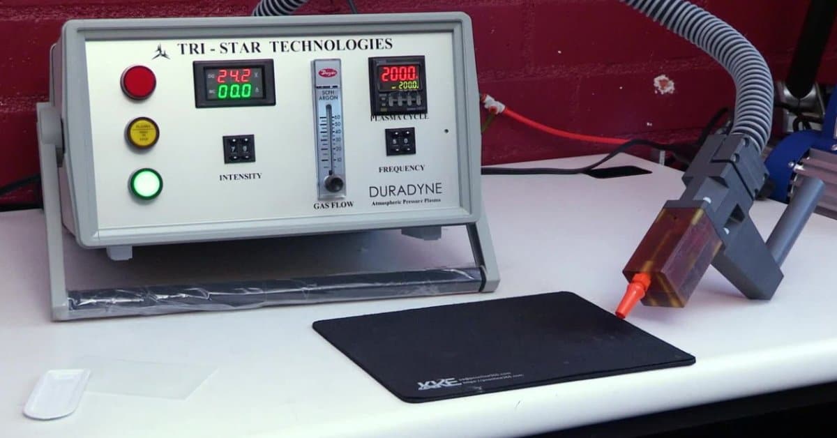 An atmospheric pressure plasma system displayed on a workbench. The setup includes a control unit with digital displays, gas flow controls, and a handheld plasma applicator positioned beside a treatment surface.