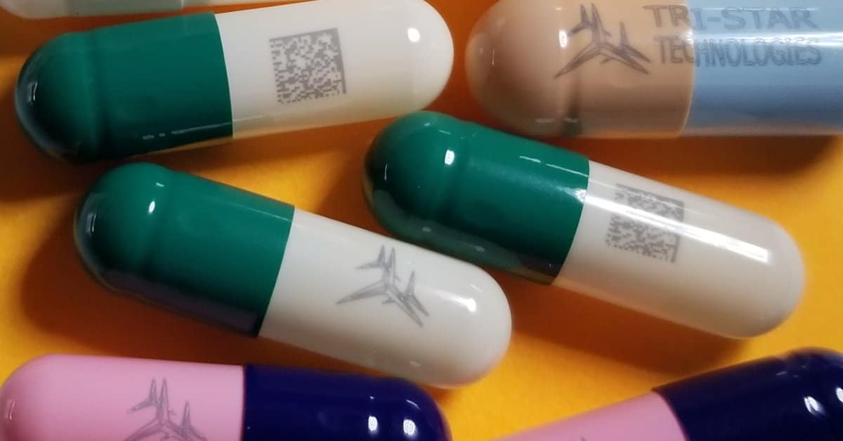 A close-up of eight oral medication capsules of various colors with various imprints, including QR codes and logos.