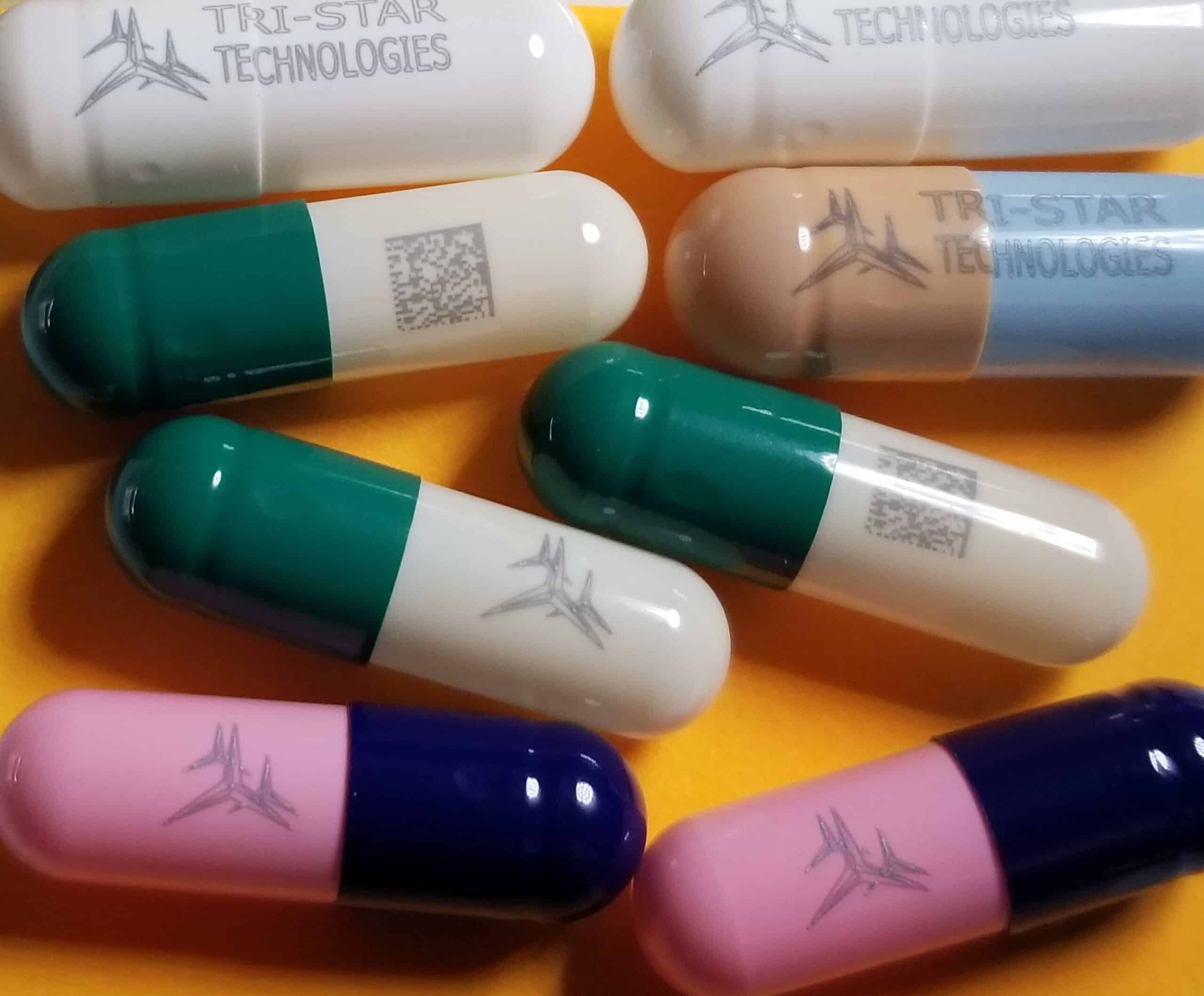 A close-up of various multi-colored prescription capsules with the Tri-Star Technologies logo and QR codes printed on them.