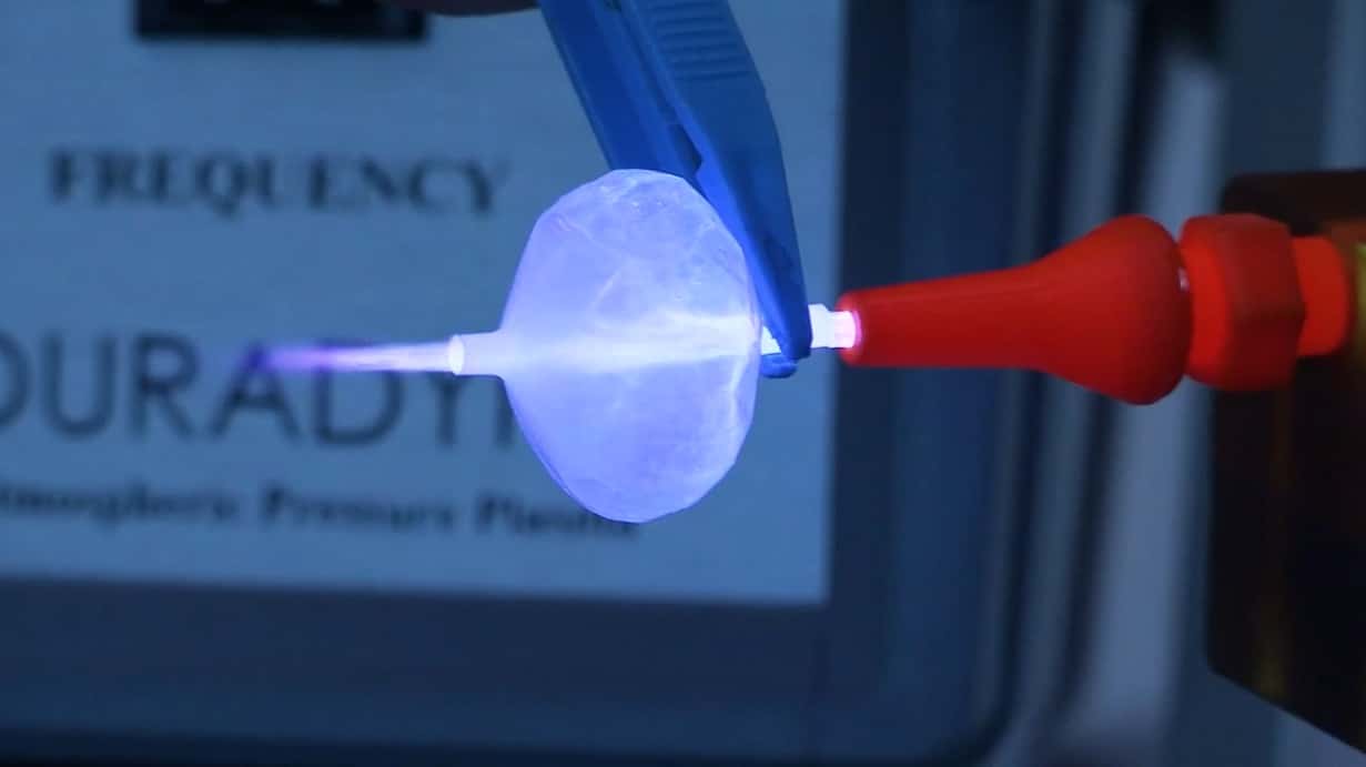 A close-up of a blue plier holding a thin, round, plastic component up to the DURADYNE plasma surface cleaning system.