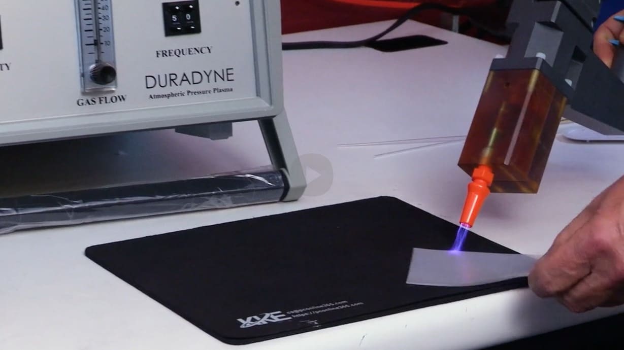 A close-up of a hand holding up a small plastic substrate to the nozzle of the DURADYNE system for plasma surface cleaning.